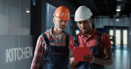 Caucasian engineer and african electric technician walking inside building hallway and inspecting light bulbs, controling electric system and coworking.
