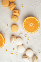 Orange and Vanilla French Macaroons with orange fruit filling on a creamy white clay background, decorated with small chamomiles and fresh sliced oranges