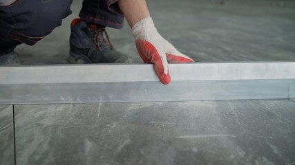 The foreman checks the level of the laid tiles on the floor. A worker in a building uniform checks...