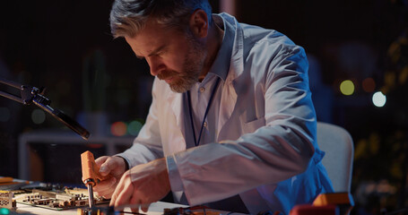 Professional adult caucasian repairman doing technical repairment circuit board soldering for broken drone device in dark office laboratory interior. Engineering,