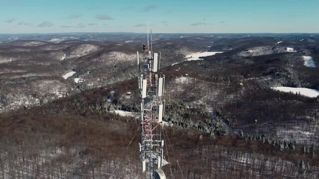 Take a look at the aerial view of 4G and 5G telecommunication tower located in hilly area covered in the snow and tall pine trees.