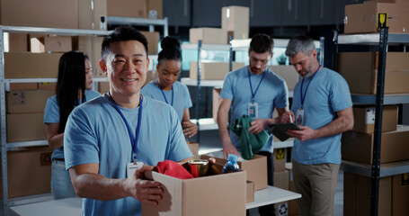 Multi-ethnic team of volunteers collaborating in workshop. Chinese male adult worker collecting food supply free delivery box smiling into camera.
