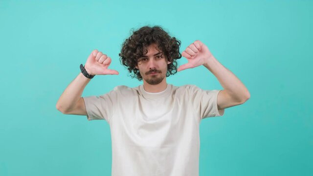Portrait of unsatisfied bearded man with curly hair making thumbs down gesture against turquoise background. Studio shot.