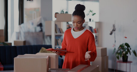 Black young attractive busy woman counting carton boxes and checking everything using digital tablet packing for moving house.