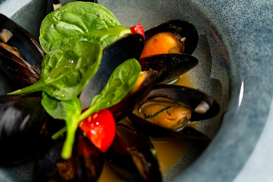 Cooked mussels in a sauce, served with spinach and cherry tomatoes on a grey plate on a black background.