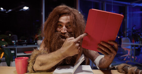 Portrait face of caveman in furry clothes yelling at camera pointing finger and showing modern...
