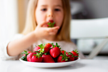 Cute little girl eating fresh strawberry in the kitchen. Healthy vitamin snack for kids. Ripe fresh berries. Harvest season. Natural vitamins .