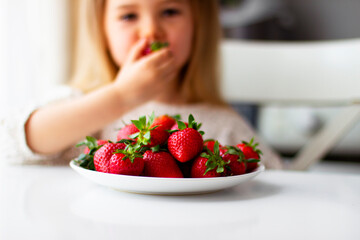 Cute little girl eating fresh strawberry in the kitchen. Healthy vitamin snack for kids. Ripe fresh berries. Harvest season. Natural vitamins .