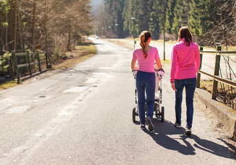 Two young woman in pink tops walking with baby carriage over asphalt road trees both sides, on sunny day, view from behind