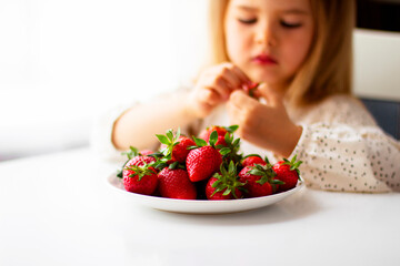 Cute little girl eating fresh strawberry in the kitchen. Healthy vitamin snack for kids. Ripe fresh berries. Harvest season. Natural vitamins .