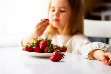 Cute little girl eating fresh strawberry in the kitchen. Healthy vitamin snack for kids. Ripe fresh berries. Harvest season. Natural vitamins .