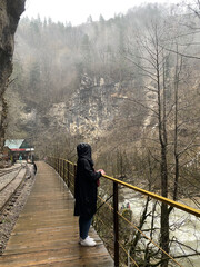 Fototapeta premium Side view of young woman standing on platform in mountainous terrain. Female tourist enjoys spectacular view of mountain landscape in rainy and cloudy weather.