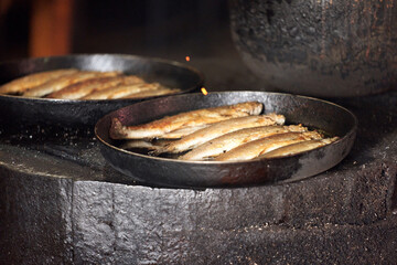  Fish in black cast-iron pans is fried on an open fire in an enclosed room. Selective focus.