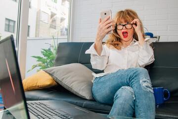 Fototapeta premium surprised woman looking at mobile phone on home sofa with computer