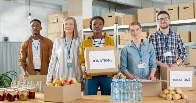 Mixed-race joyful young and senior charity organization male and female workers working at charity storage looking at camera with smile on face. Donation box, volunteer center, volunteering concept