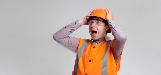 young engineer architect clutching his head in helmet on grey studio background, foreman shocked