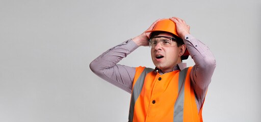 young engineer architect clutching his head in helmet on grey studio background, foreman shocked