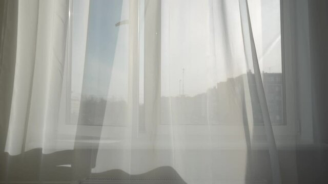 Transparent curtain on open window moved by the wind at sunny day, sun rays shine through the transparent tulle on window of apartment room. High quality 4k footage