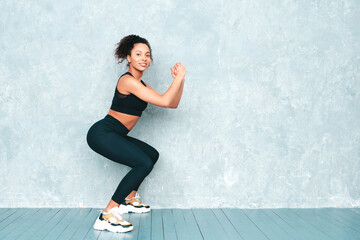 Fototapeta premium Fitness smiling black woman in sports clothing with afro curls hairstyle.She doing squats. Young beautiful model with perfect tanned body.Female in studio near gray wall