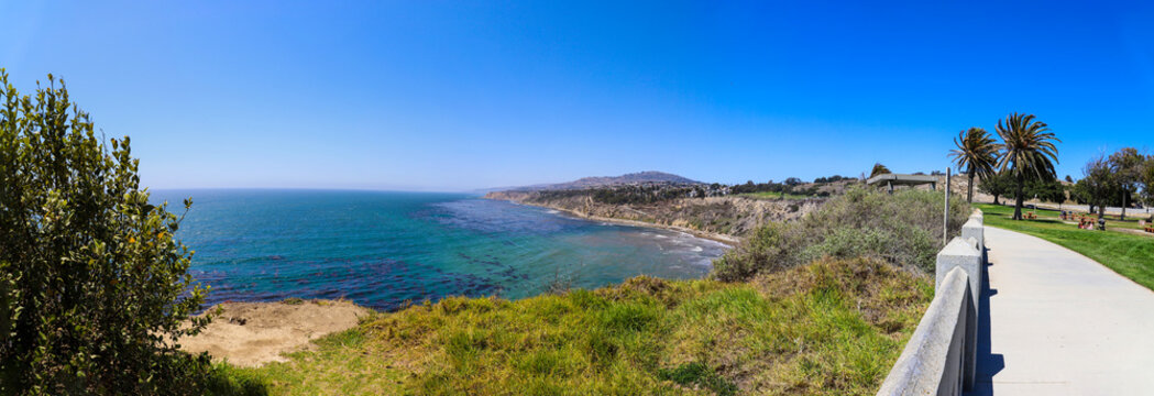 a panoramic shot of the coast with vast deep blue ocean water and waves crashing into the beach with blue sky and lush green trees and plants on the hillside at Point Fermin Park in San Pedro CA