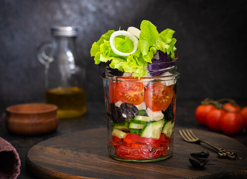 Healthy greek salad with fresh vegetables and feta cheese in a glass jar on a dark concrete background. Salad recipes.