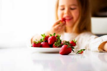  Cute little girl eating fresh strawberry in the kitchen. Healthy vitamin snack for kids. Ripe fresh berries. Harvest season. Natural vitamins .