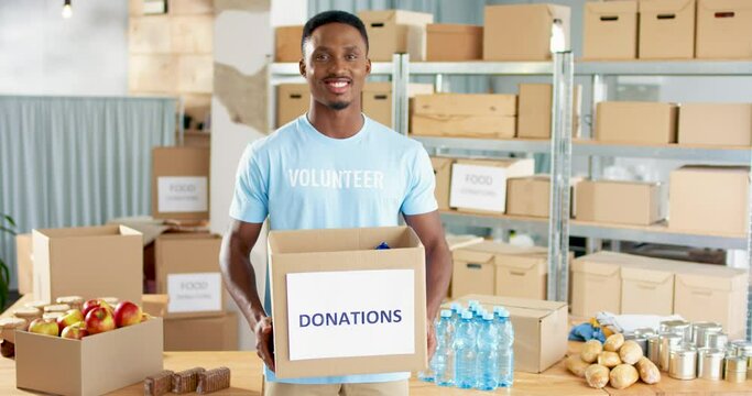 Portrait of young African American handsome cheerful guy volunteer standing in warehouse holding box with donations and social help in hands, looking at camera and smiling, charity center concept