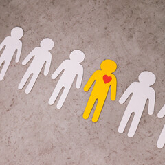 Cut out of white paper figures of people in line. In center is yellow man, with red heart in chest....