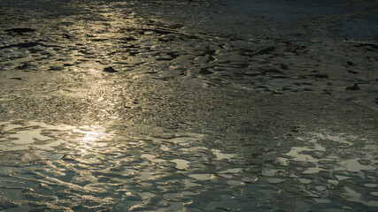 bumpy surface melting wet ice in the thaw in early spring with numerous shallow puddles and reflection of the morning or evening sun
