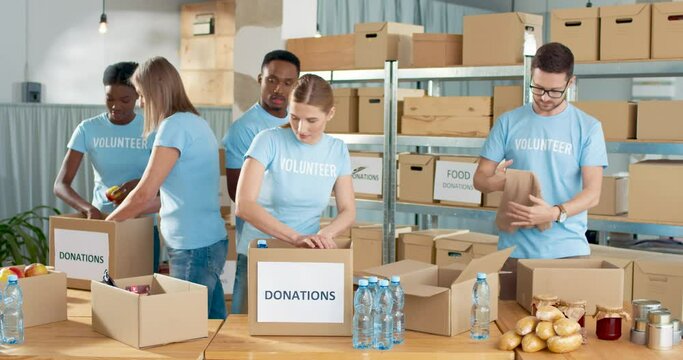 Mixed-race joyful positive male and female volunteers working in community center preparing and packing social help box, man making check list of donations box, volunteering and donating concept