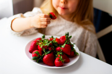 Cute little girl eating fresh strawberry in the kitchen. Healthy vitamin snack for kids. Ripe fresh berries. Harvest season. Natural vitamins .