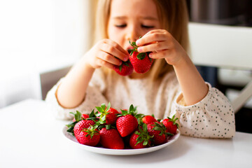 Cute little girl eating fresh strawberry in the kitchen. Healthy vitamin snack for kids. Ripe fresh berries. Harvest season. Natural vitamins .