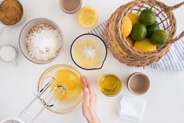 Woman hands about to mix some eggs with a blender in a bowl on a white table full of fresh muffin ingredients.
