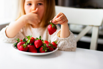 Cute little girl eating fresh strawberry in the kitchen. Healthy vitamin snack for kids. Ripe fresh berries. Harvest season. Natural vitamins .
