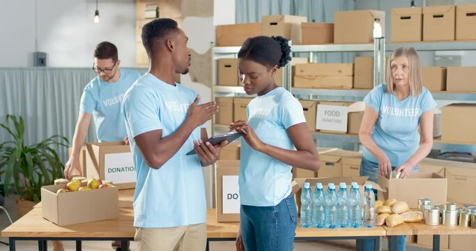 African American young happy male and female volunteers standing in warehouse typing browsing online on tablet device and speaking. Caucasian workers packing donations help in box, volunteering