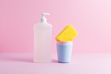 Creative still life with dishwashing supplies. White bottle of dishwash liquid and yellow kitchen cleaning sponge in stack of plastic cups. Front view, copy space, pink background.