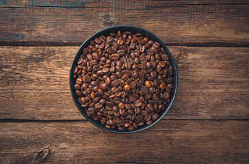 Coffee beans in a black round ceramic plate on a brown wooden background.