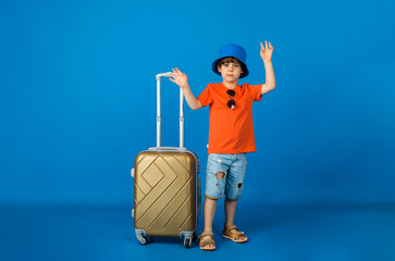 caucasian little boy traveler in panama holds a yellow suitcase on a blue background with space for text