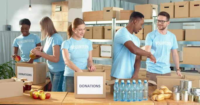 Mixed-race happy positive male and female different volunteers working in charity organization center preparing and packing social help box, donations box, volunteering and donating concept