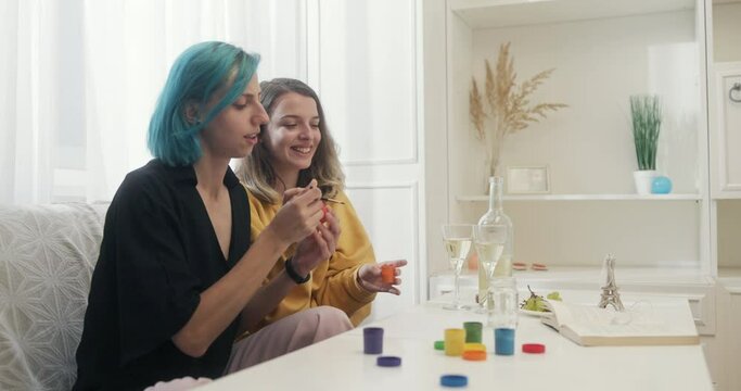 Two young lesbian girls sitting on sofa at home in a bright cozy apartment painting with paints creatively spending time together smiling and having fun