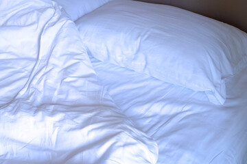Unmade bed of clean and white pillow, blanket, and bed sheet with morning sunshine