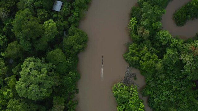 Aerial top view of a wooden canoe with motor driving over a broad tropical river in a rainforest towards a small indigenous community in the Amazon of Ecuador