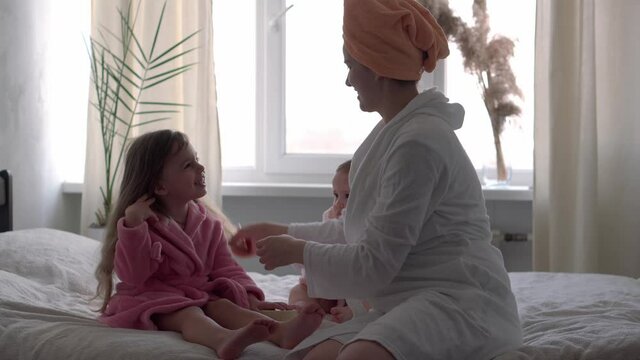 Authentic cute young woman combing long hair of baby girl. mom with 2 children daughters different ages in bathrobes after bath on white bed oposite window. Spa, motherhood, childhood, family concept.