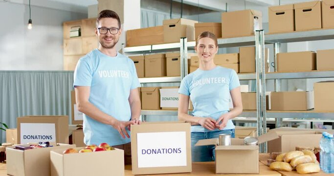 Caucasian young joyful couple of man and woman charity workers putting food products in box standing in warehouse looking at camera and smiling. Volunteers prepare donations. volunteering and donating