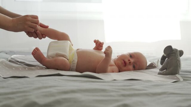 Mother with baby boy doing exercise simple exercises to make baby bones and muscles stronger, newborn infant lying on bed against the window. High quality 4k footage