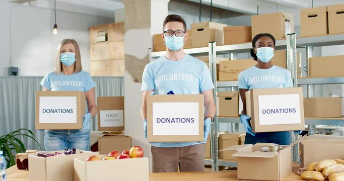Mixed-race nice male and female volunteers standing in charity organization warehouse holding donations boxes in hands. Volunteer activity, shipping delivery charitable stock, social help concept
