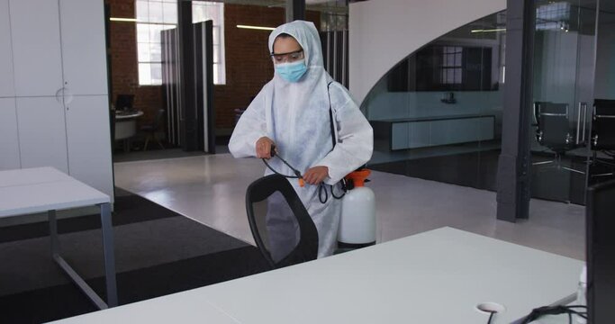 Female cleaner wearing protective face mask and overalls disinfecting office and furniture