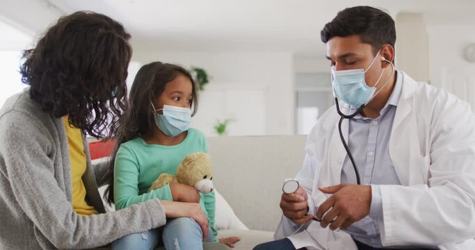 Hispanic Male Doctor Holding Stethoscope Talking To Mother And Daughter At Home Wearing Face Masks