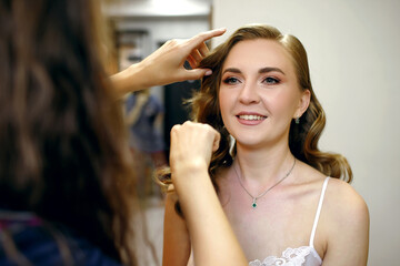 Happy bride doing her hair in a beauty salon getting ready for the wedding day. Styling for a beautiful young woman is done by a professional master stylist. Preparing for a wedding ceremony or party