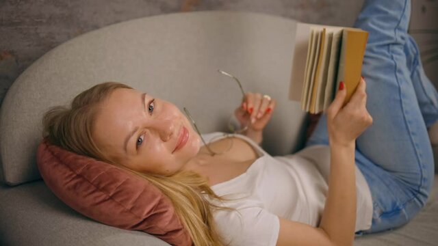 Young woman holding book lying on sofa and looking at you orbit shot